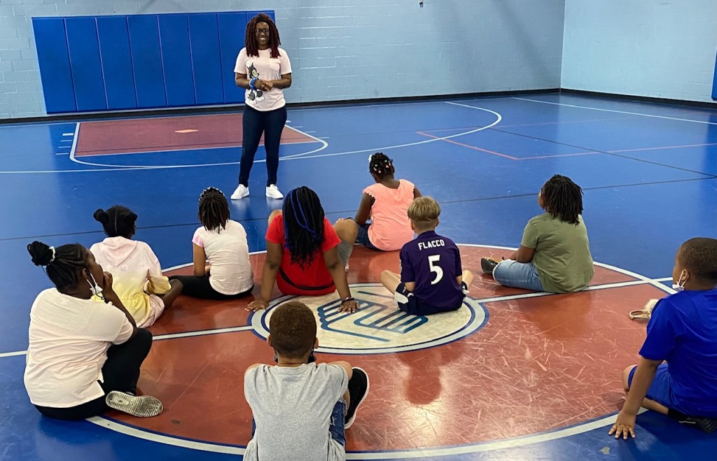 Nine young boys and girls, sitting in a gym, listening to an adult wearing a pink tom and jerry shirt.